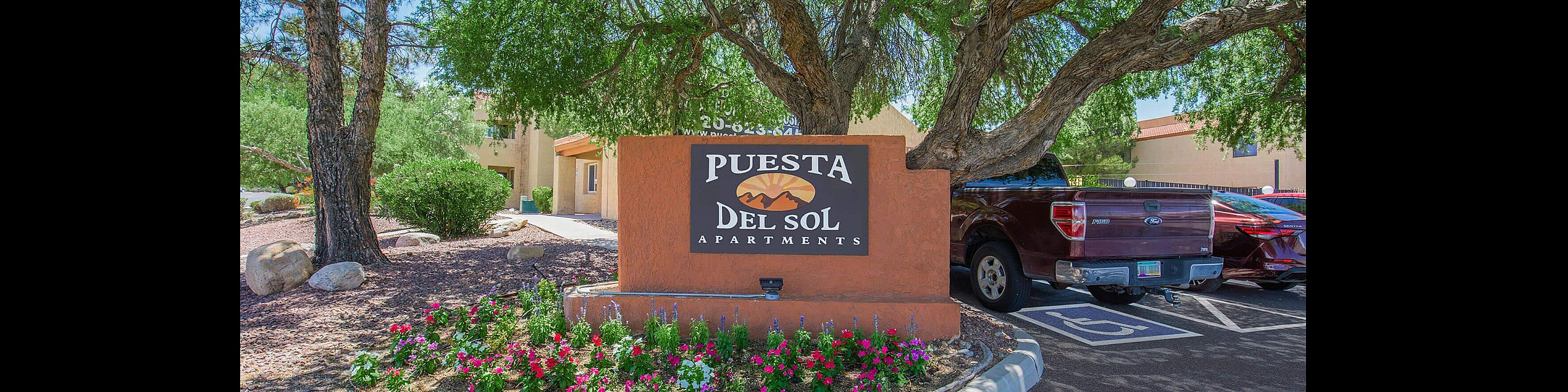 Inviting entrance to Puesta Del Sol Apartments, featuring vibrant landscaping and shaded by lush green trees.