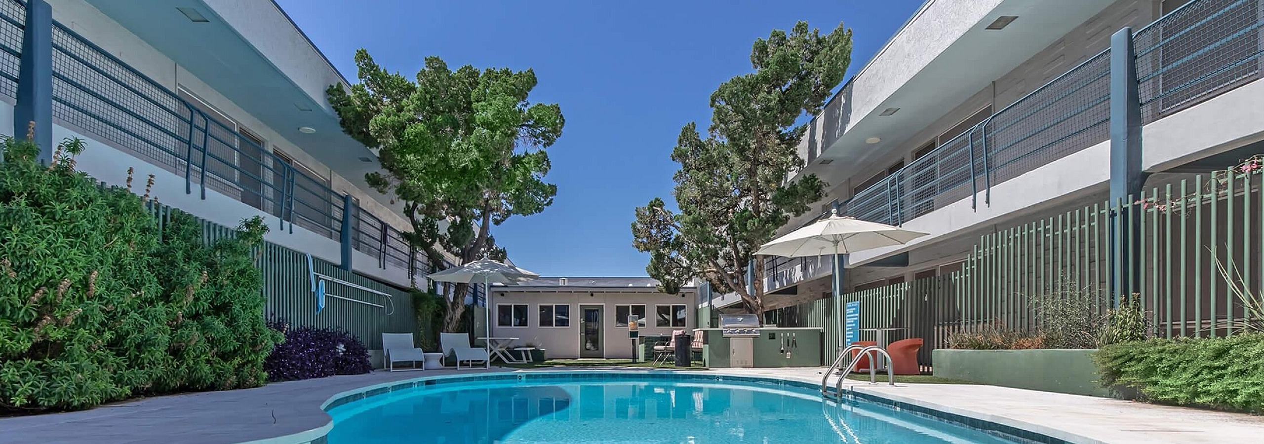 Inviting apartment complex with a sparkling outdoor pool, stylish lounge chairs, and vibrant landscaping under a clear blue sky.