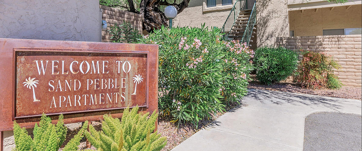 Inviting entrance to Sand Pebble Apartments with lush greenery and charming desert landscaping.