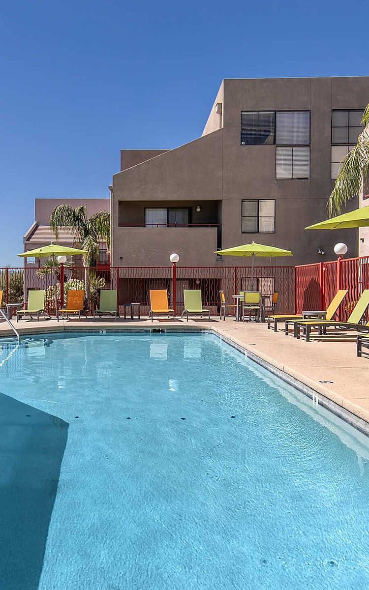 Inviting apartment pool area with vibrant lounge chairs, umbrellas, and clear blue water under a bright sky.