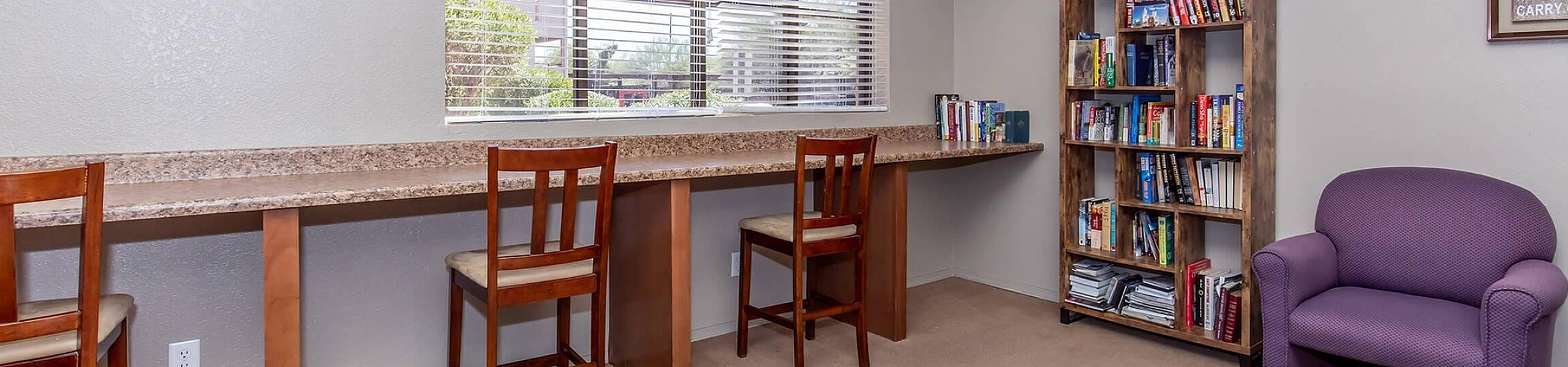 Bright and inviting study area featuring a long desk with chairs, ample natural light from large windows, and a cozy reading nook with a bookshelf and armchair.