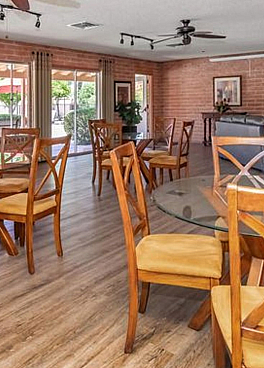 Bright and spacious community room with elegant wooden furniture, ceiling fans, and large windows offering a view of the pool area.