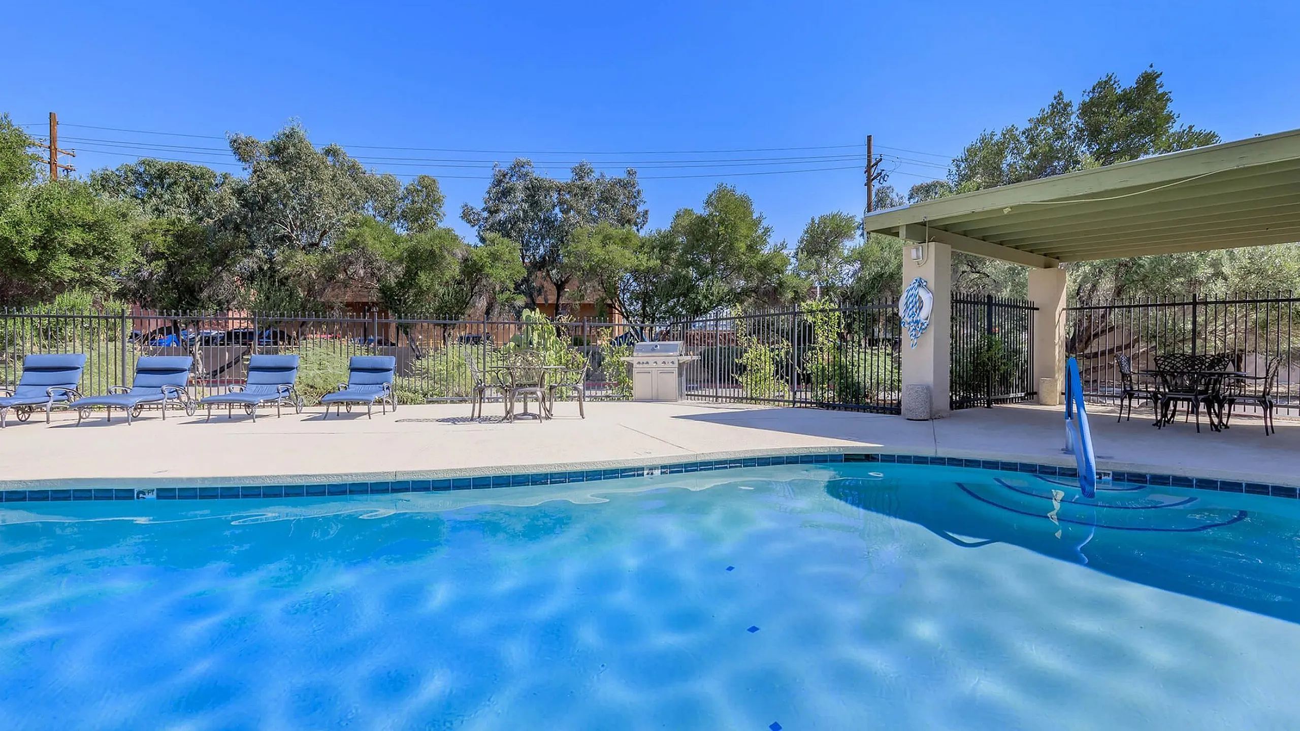 Inviting outdoor pool area with lounge chairs and shaded seating, surrounded by lush greenery and blue skies.