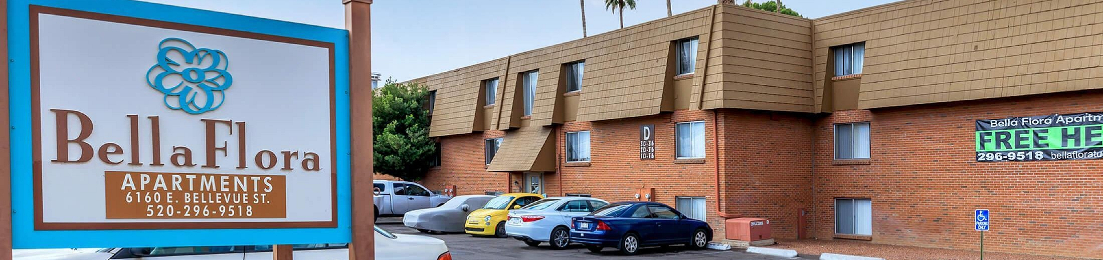 Inviting exterior view of Bella Flora Apartments with vibrant flower beds and ample parking space under sunny skies.