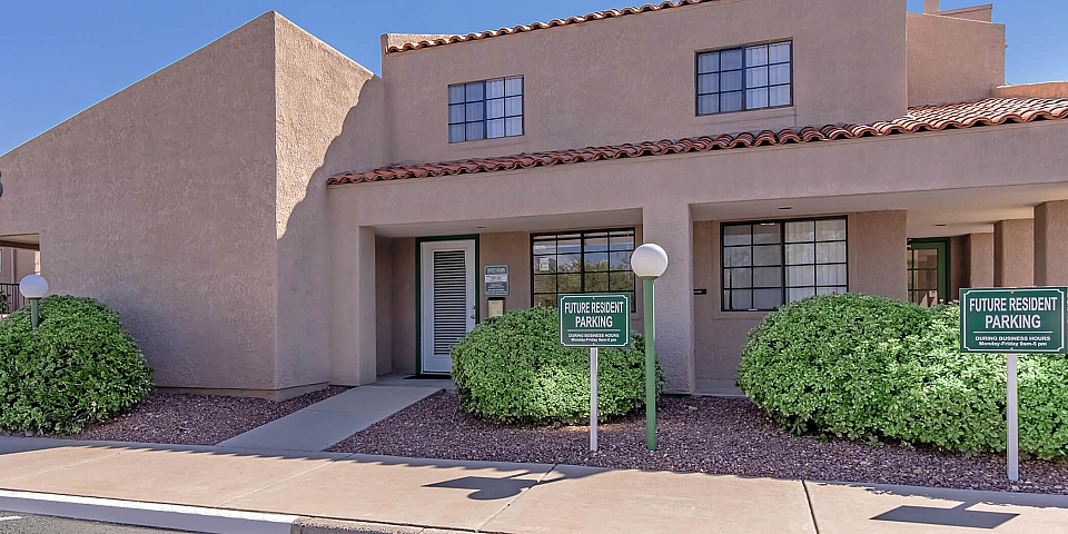 Charming apartment complex exterior with adobe-style architecture, parking spaces for future residents, and lush landscaping under clear blue skies.