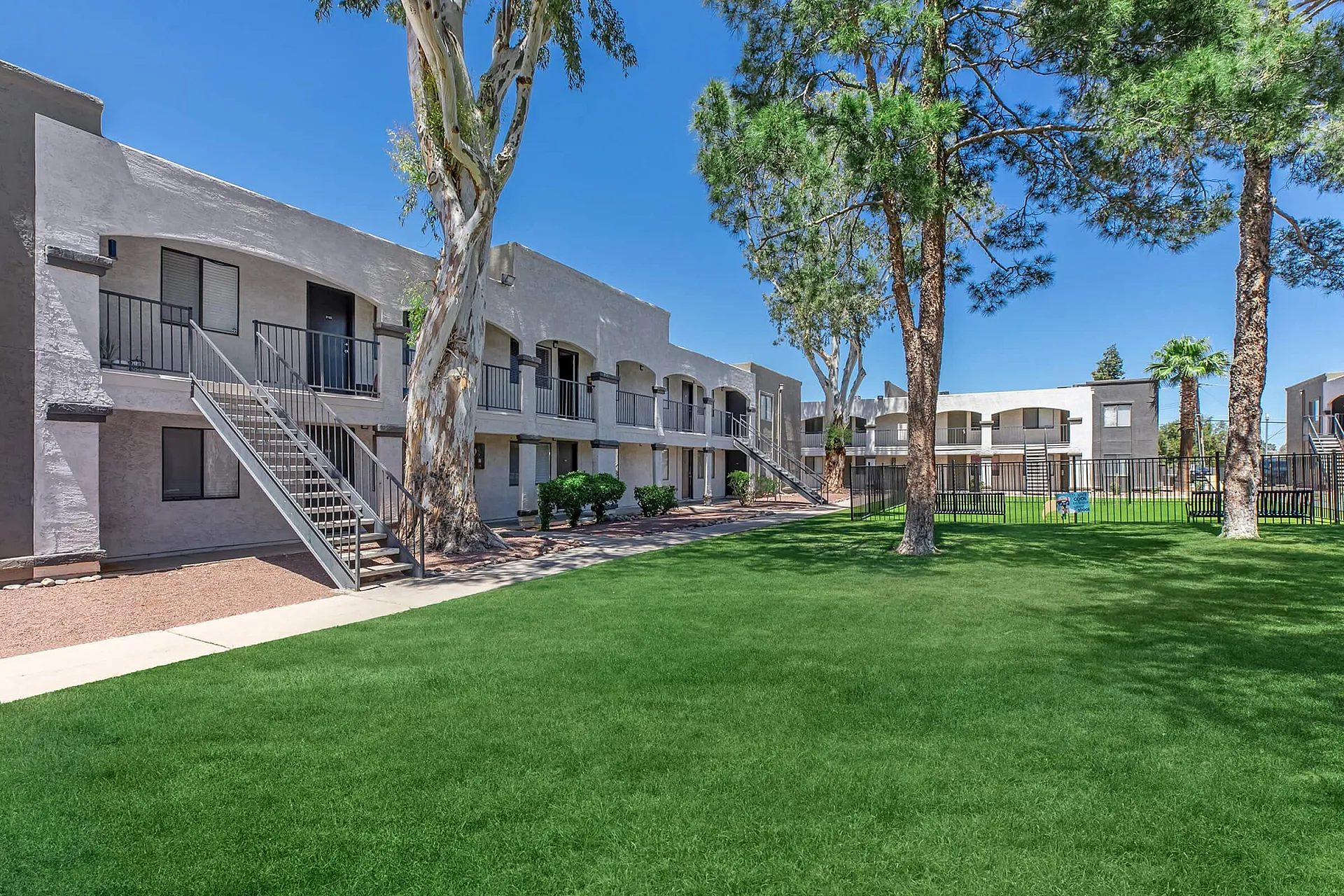 Beautiful apartment complex with lush green grass, large trees, and modern two-story buildings under a clear blue sky.