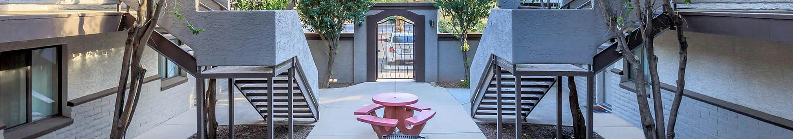 Elegant apartment courtyard featuring lush trees, dual staircases, and a cozy picnic area under a clear blue sky.