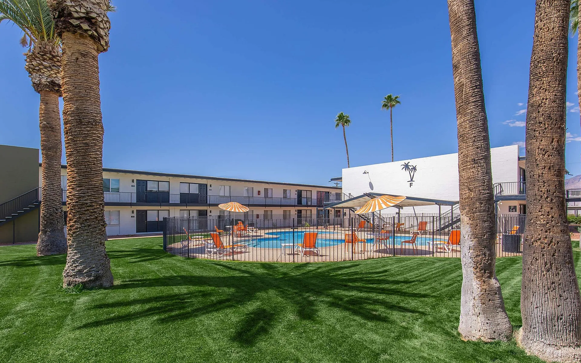 Inviting apartment complex featuring a lush green lawn, palm trees, and a sparkling pool with vibrant orange lounge chairs and umbrellas.