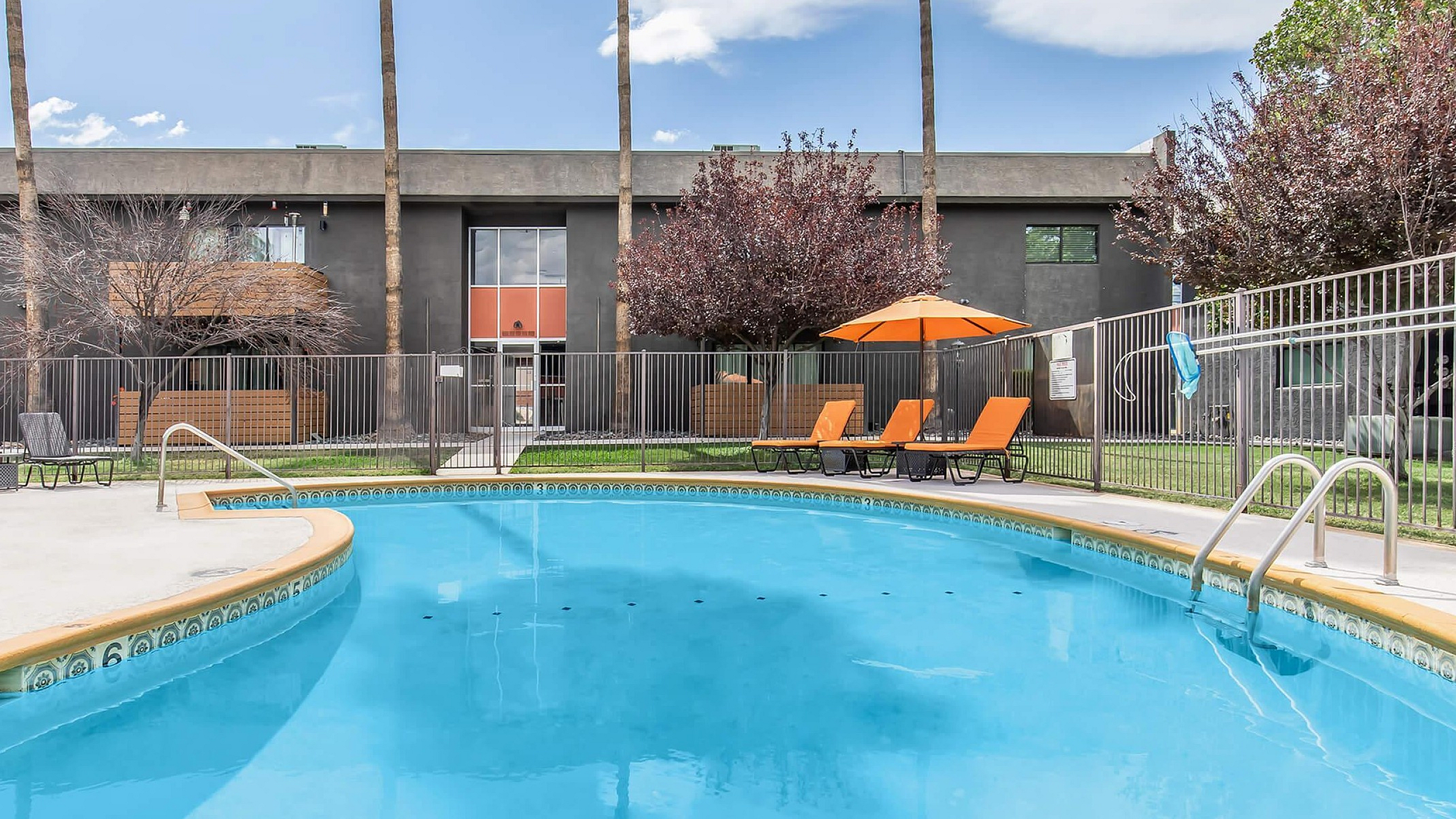Bright and inviting outdoor pool area with lounge chairs and umbrellas, surrounded by a modern apartment building and lush greenery.
