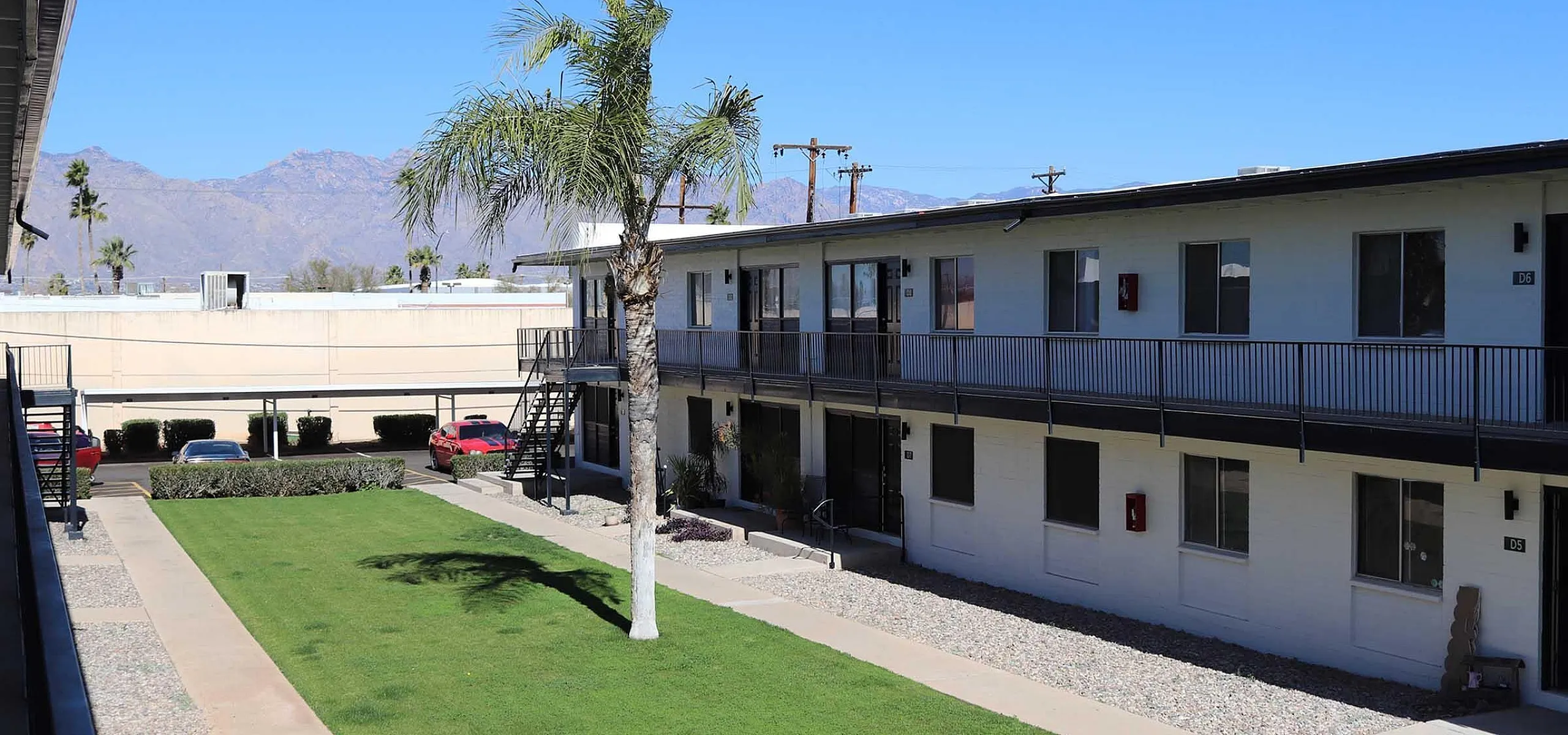 Charming apartment complex with lush green courtyard, picturesque palm trees, and stunning mountain views under a clear blue sky.