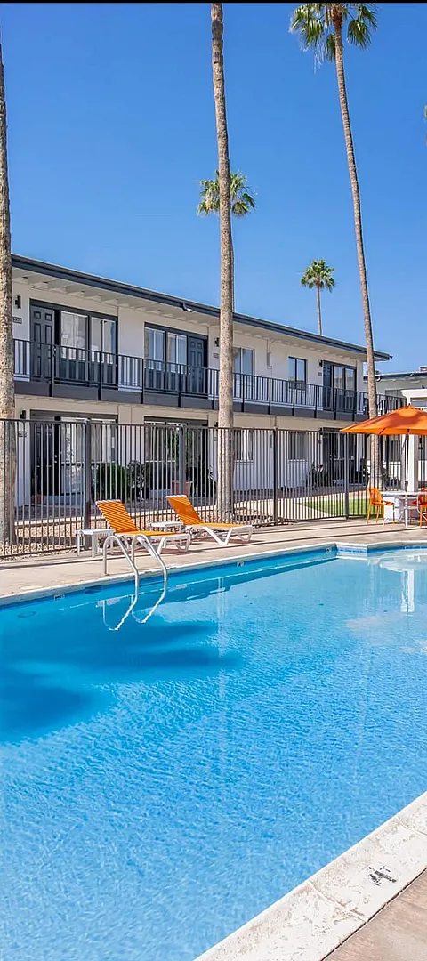 Inviting pool area surrounded by vibrant orange lounge chairs and umbrellas, set against a backdrop of elegant apartments and tall palm trees, perfect for relaxation and leisure.