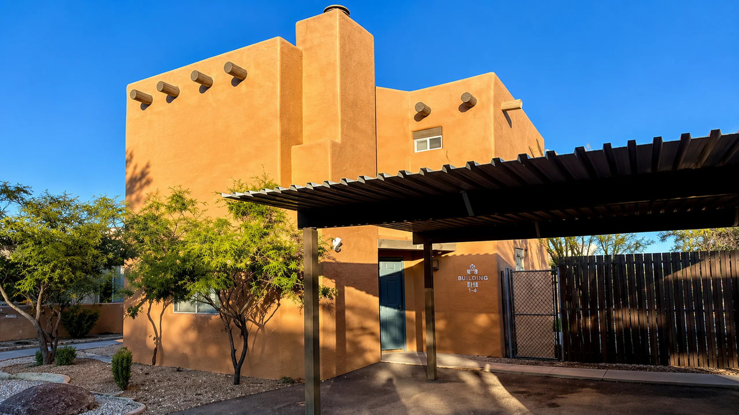 Elegant adobe-style building with a warm terracotta facade, shaded carport, and lush landscaping under a clear blue sky.