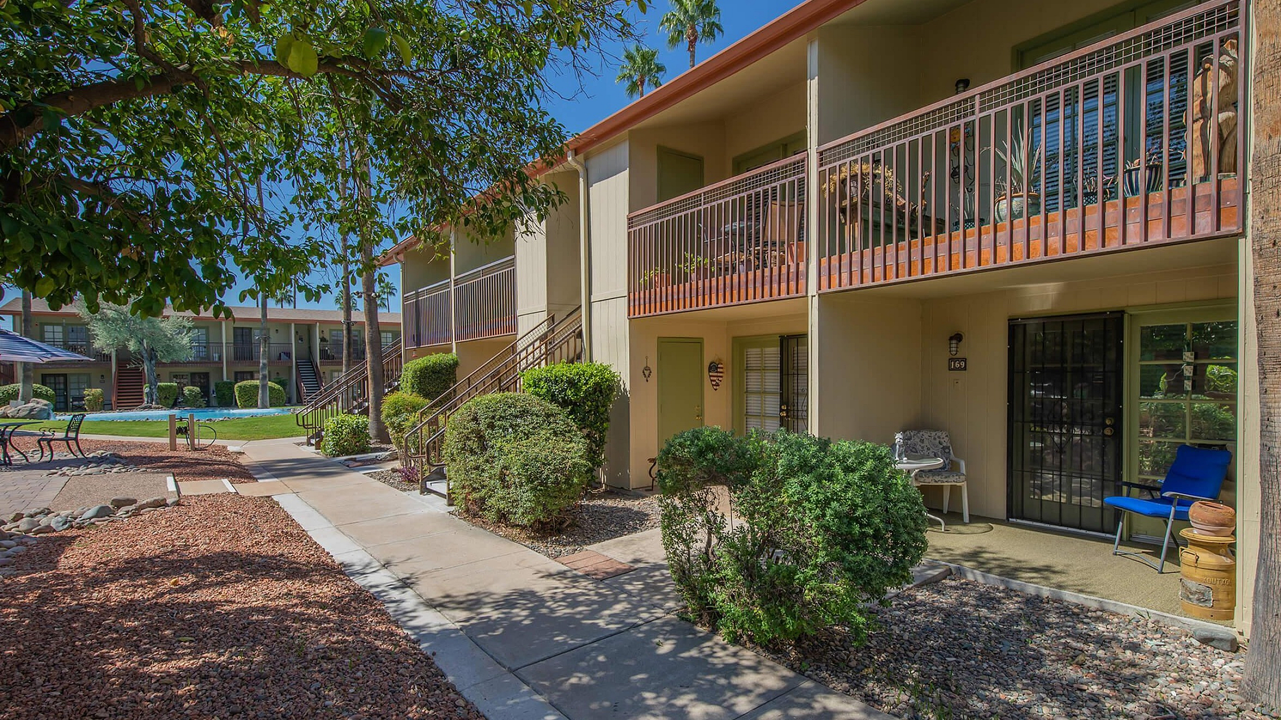 Charming apartment complex with lush landscaping, private balconies, and sunny outdoor seating under a vibrant blue sky.