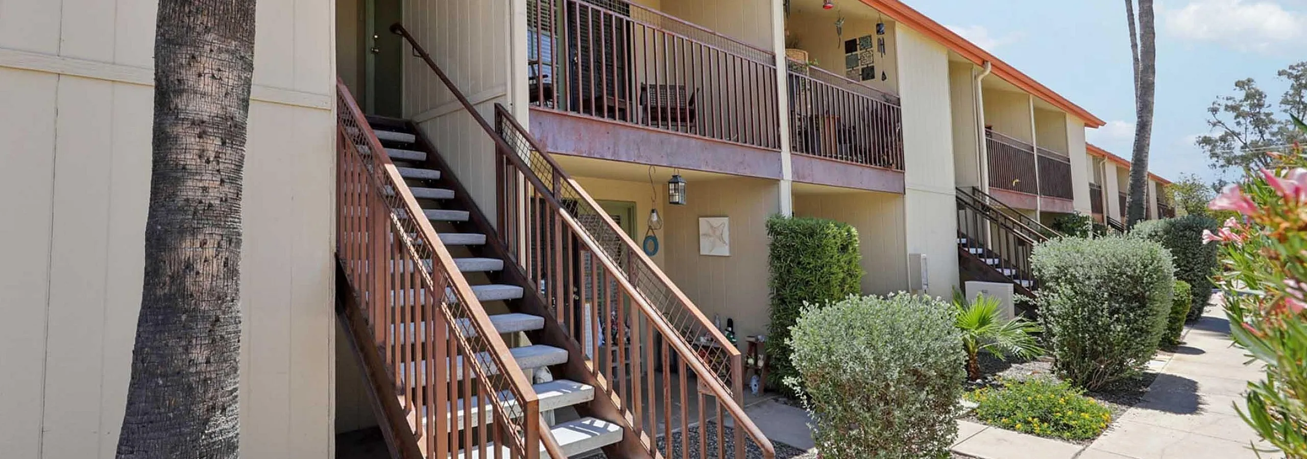 Charming apartment exterior with lush landscaping, inviting balcony views, and a warm, welcoming staircase entrance.