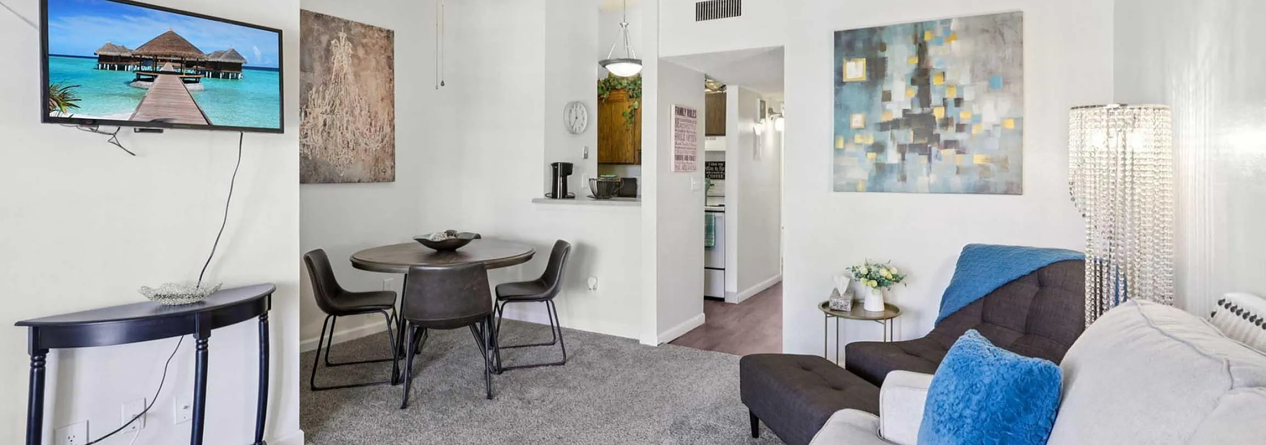 Modern living room with stylish decor, featuring a cozy sofa, vibrant artwork, and a view of the kitchen. A sleek dining table set for contemporary living.