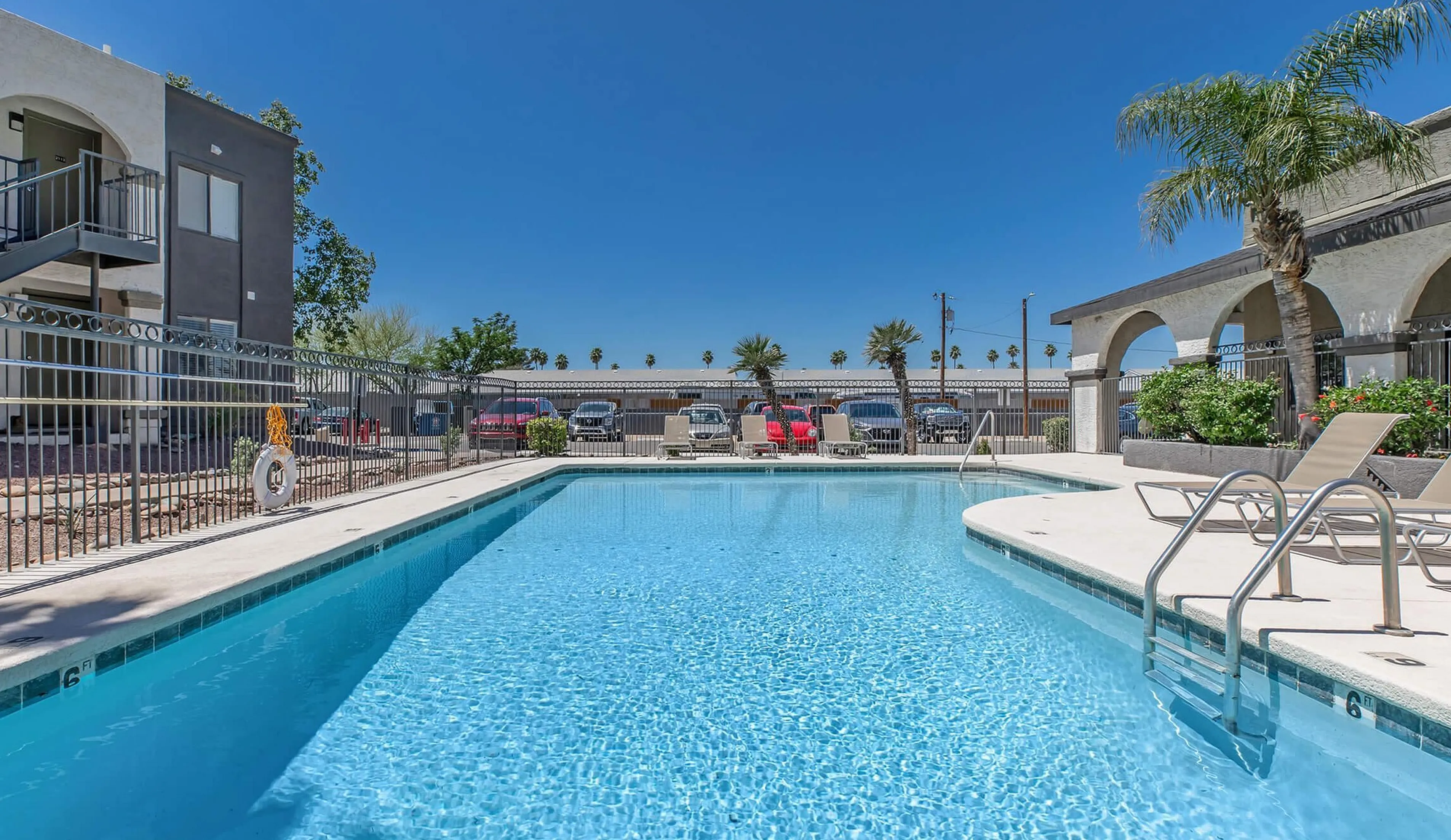 Inviting apartment complex pool area with crystal-clear water, sun loungers, and lush palm trees under a bright blue sky.