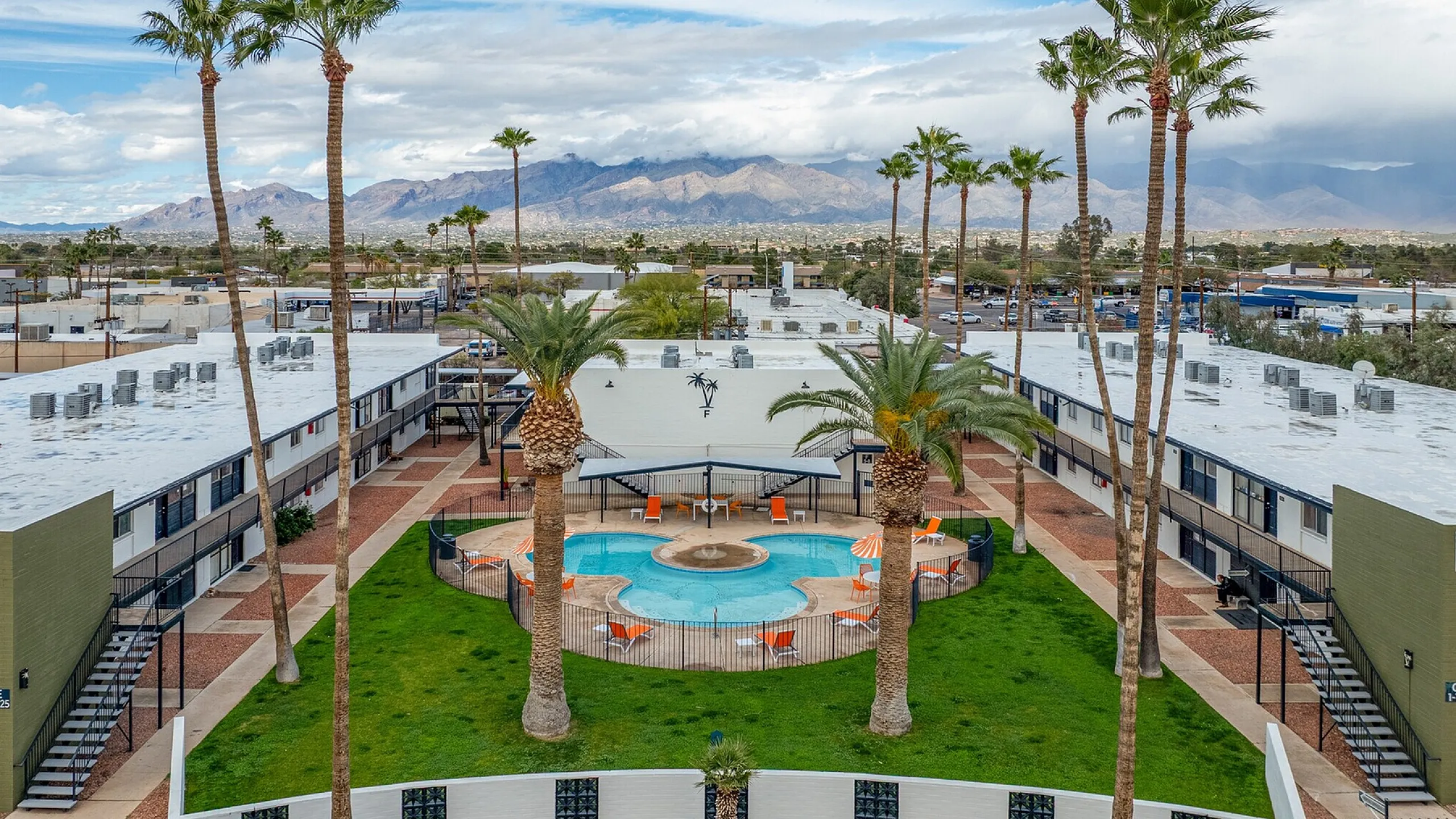 Vibrant apartment complex with a lush central courtyard and inviting swimming pool, framed by tall palm trees under a stunning mountain backdrop.