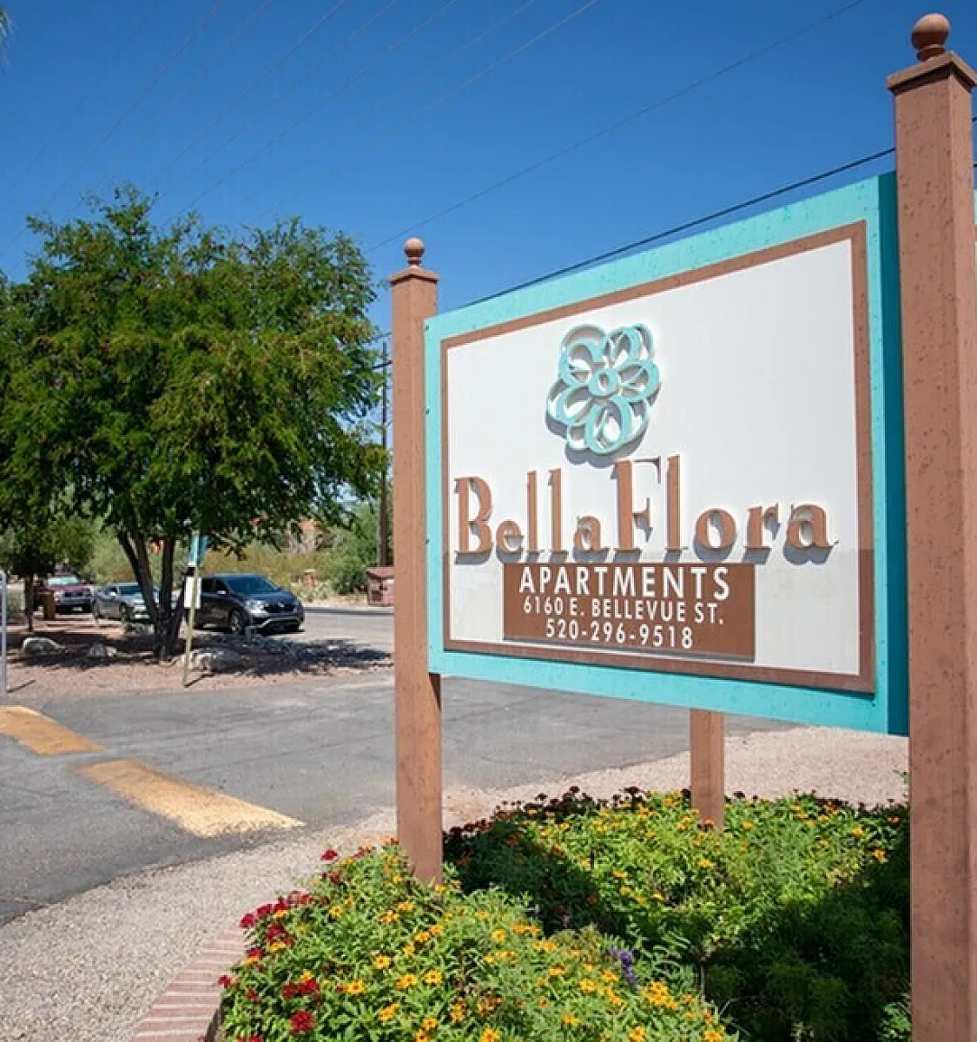 Inviting Bella Flora Apartments entrance sign surrounded by vibrant landscaping under a clear blue sky, offering a warm welcome to residents.