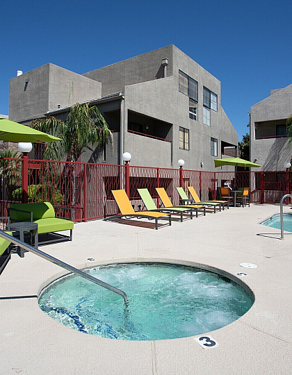 Inviting apartment complex courtyard featuring a sparkling pool, relaxing hot tub, vibrant green and orange lounge chairs, and lush palm trees under a clear blue sky.