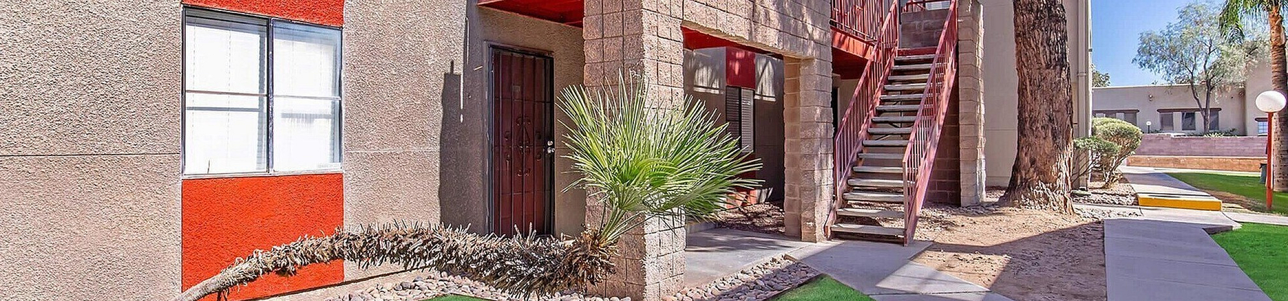 Charming apartment exterior with vibrant red accents, lush green lawns, and a welcoming staircase under a tranquil blue sky.