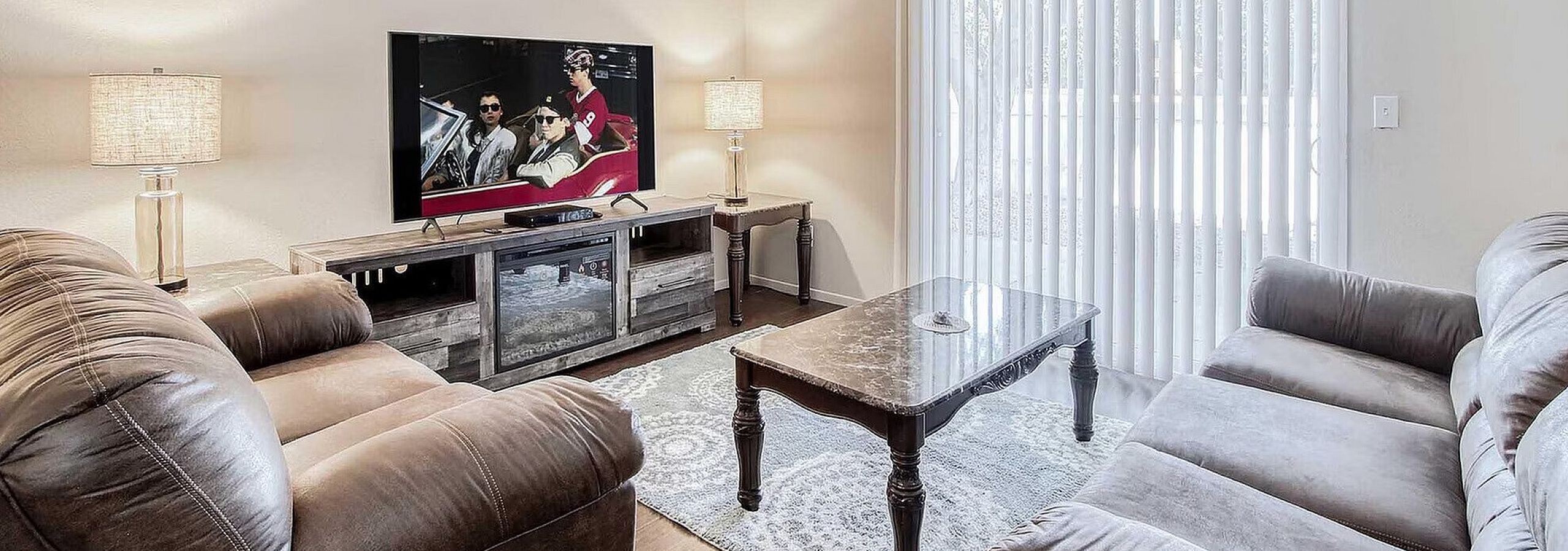 Inviting living room with plush leather sofas, elegant coffee table, and large flat-screen TV, brightly lit by natural light through vertical blinds.