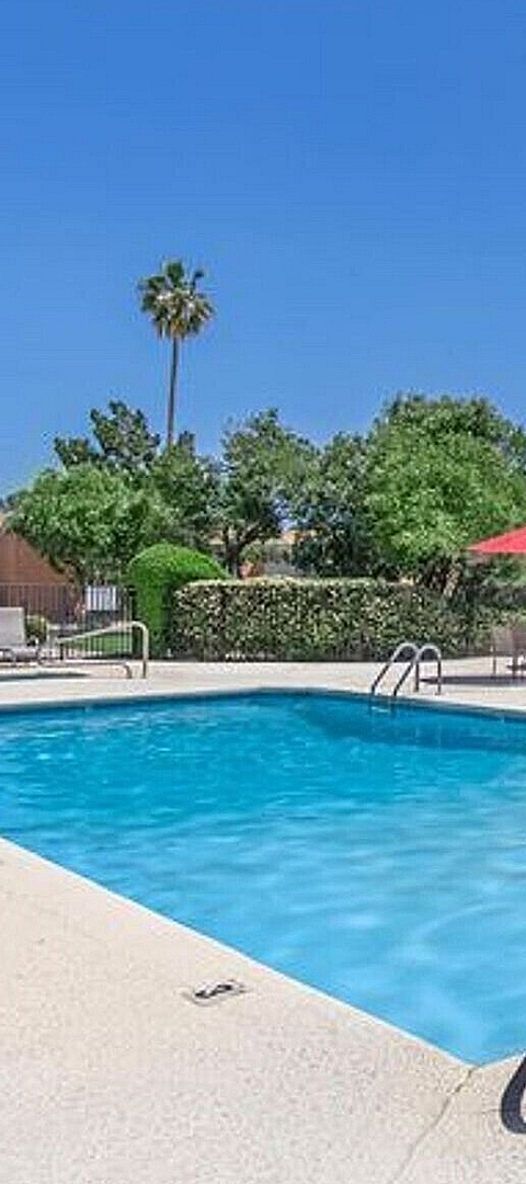 Inviting outdoor pool with comfortable lounging area and vibrant red umbrellas, surrounded by lush greenery and a charming community building under a clear blue sky.
