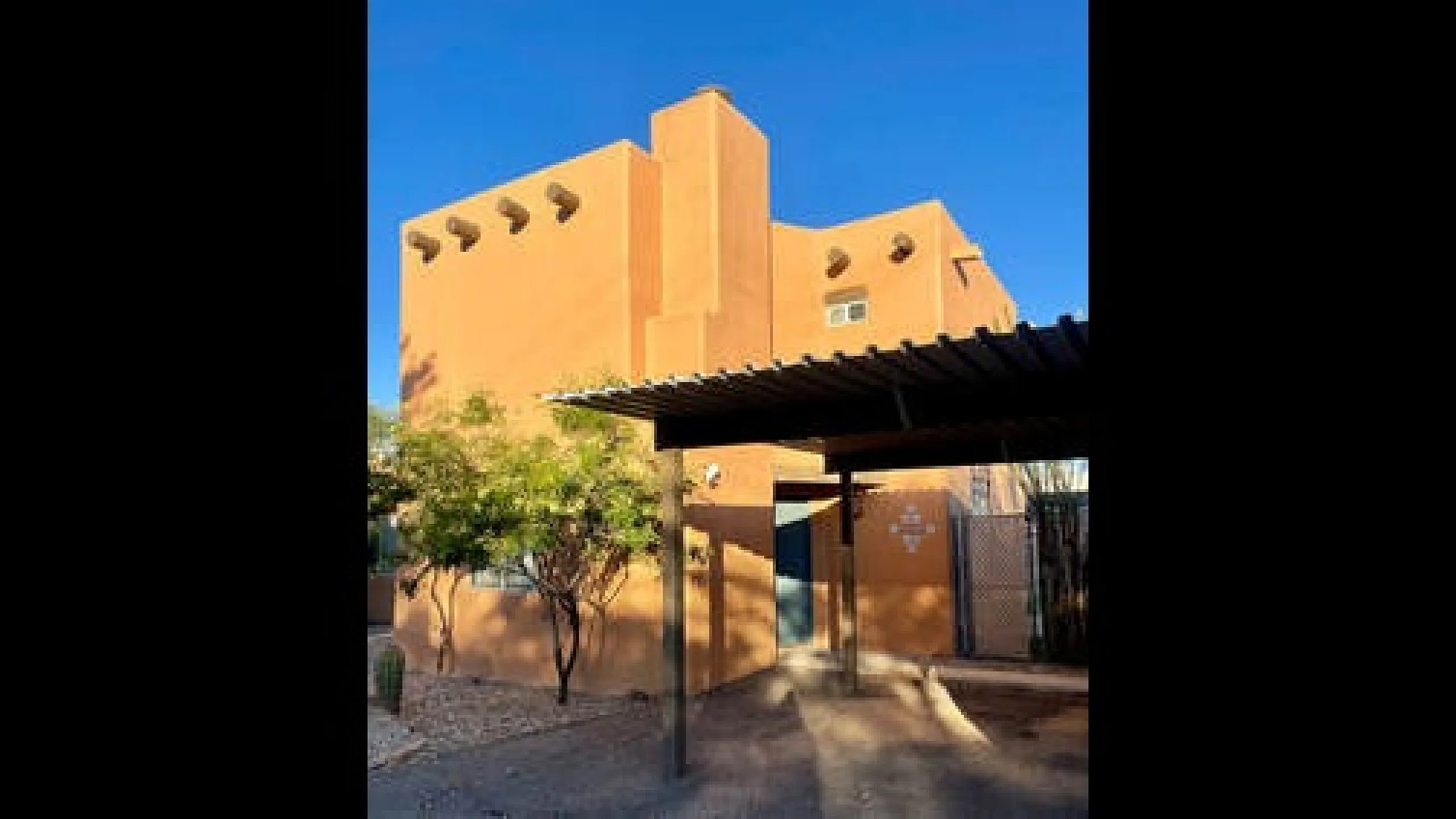 Charming adobe-style home with vibrant orange exterior, shaded carport, and desert landscaping under a clear blue sky.