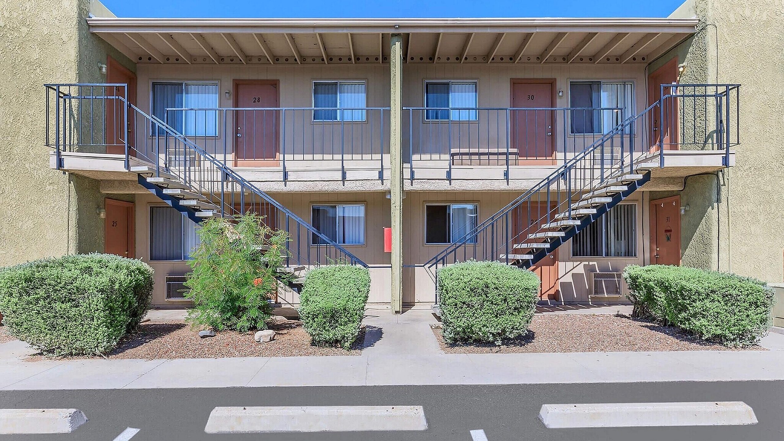 Charming two-story apartment building with modern railings, lush greenery, and freshly painted doors, offering comfortable and stylish living spaces.