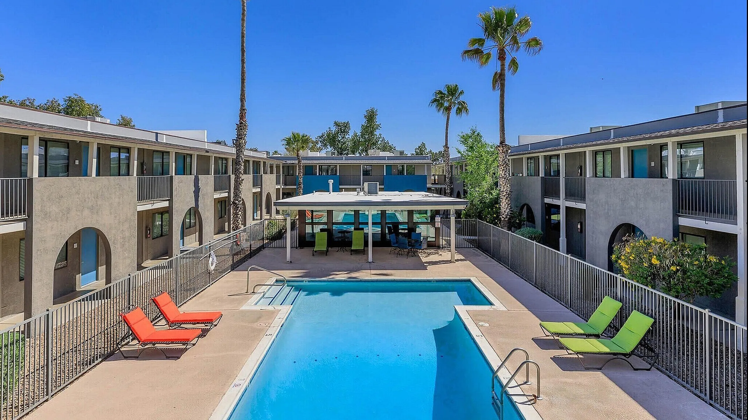 Contemporary apartment complex with a sparkling outdoor pool, vibrant lounge chairs, and lush palm trees under a clear blue sky.