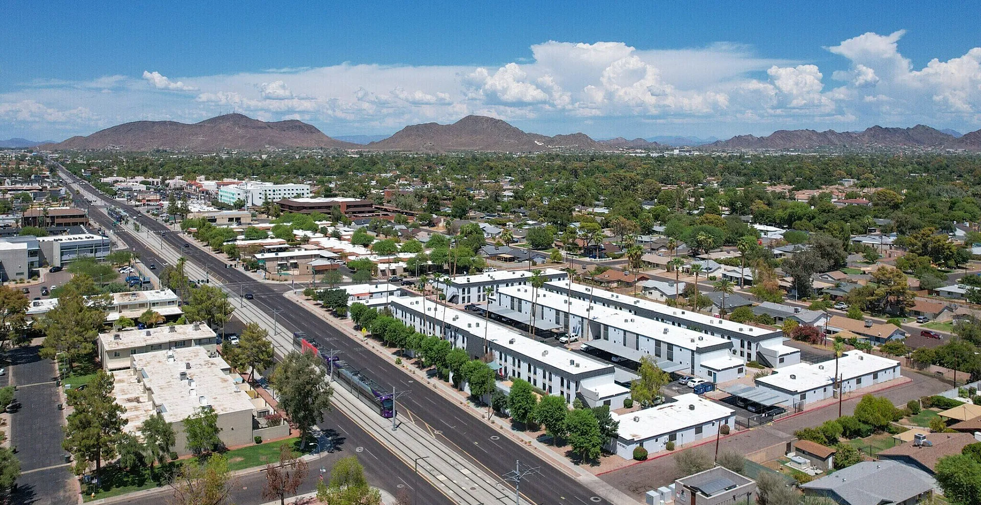 Vibrant aerial view of modern apartment complex with striking mountain backdrop, situated in a lively urban area with lush greenery and convenient road access.