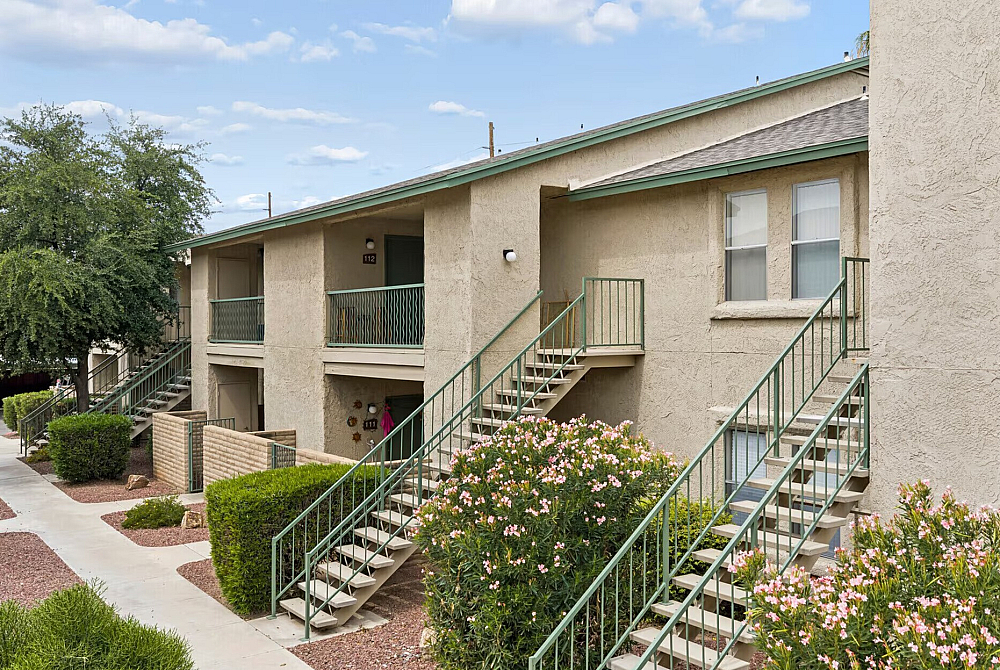 Charming two-story apartment building with green railings, surrounded by lush landscaping and vibrant flowers under a blue sky.