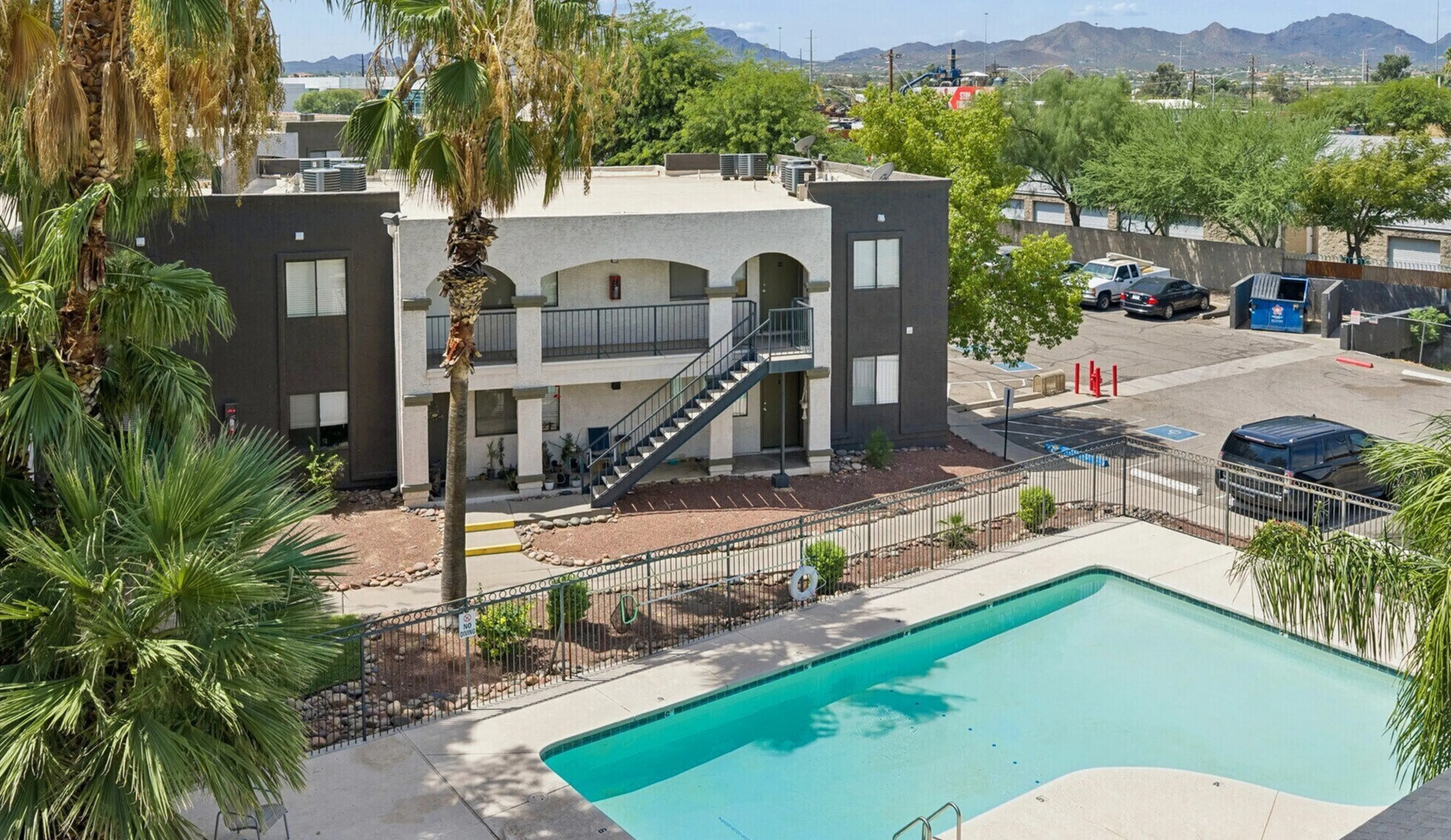 Charming apartment complex featuring a sunlit outdoor swimming pool surrounded by lush palm trees and mountain views.