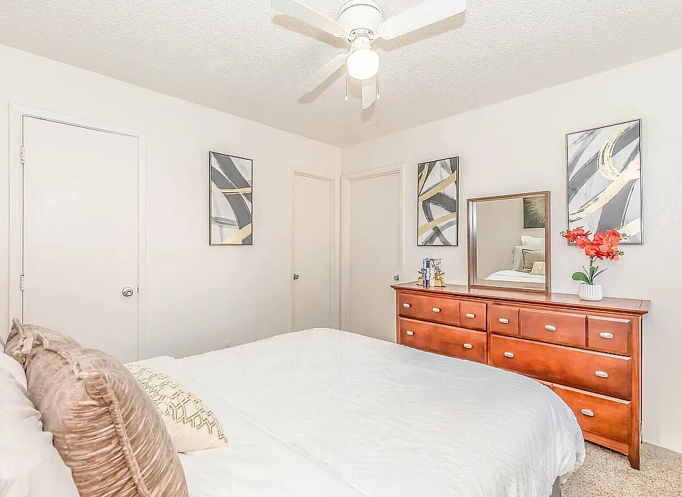 Bright and inviting bedroom with modern art, a sleek ceiling fan, and a stylish wooden dresser topped with vibrant decor.