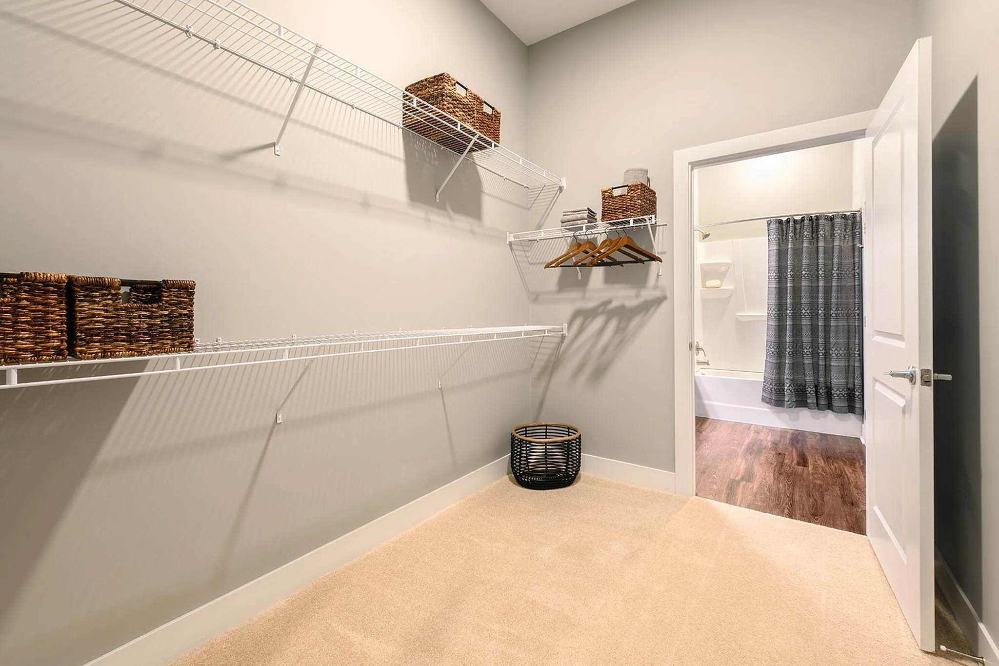 Spacious walk-in closet with ample shelving and elegant wicker baskets, leading into a modern bathroom with chic shower curtains.
