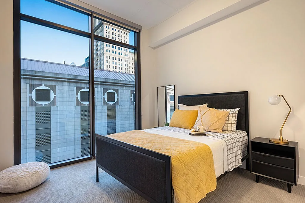 Elegant bedroom with large windows showcasing a stunning city view, complete with a cozy bed adorned with stylish yellow accents.