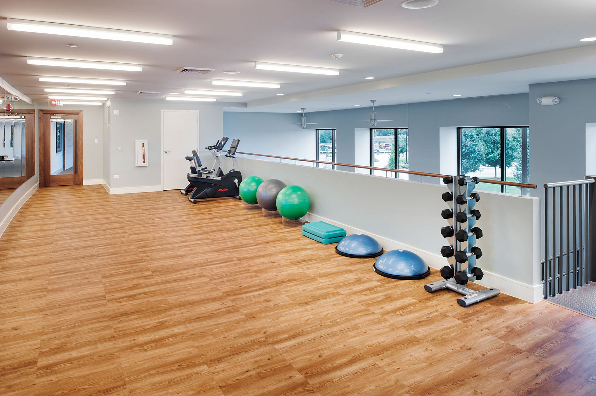 Bright and spacious fitness room with modern exercise equipment, wooden flooring, and large windows inviting natural light.