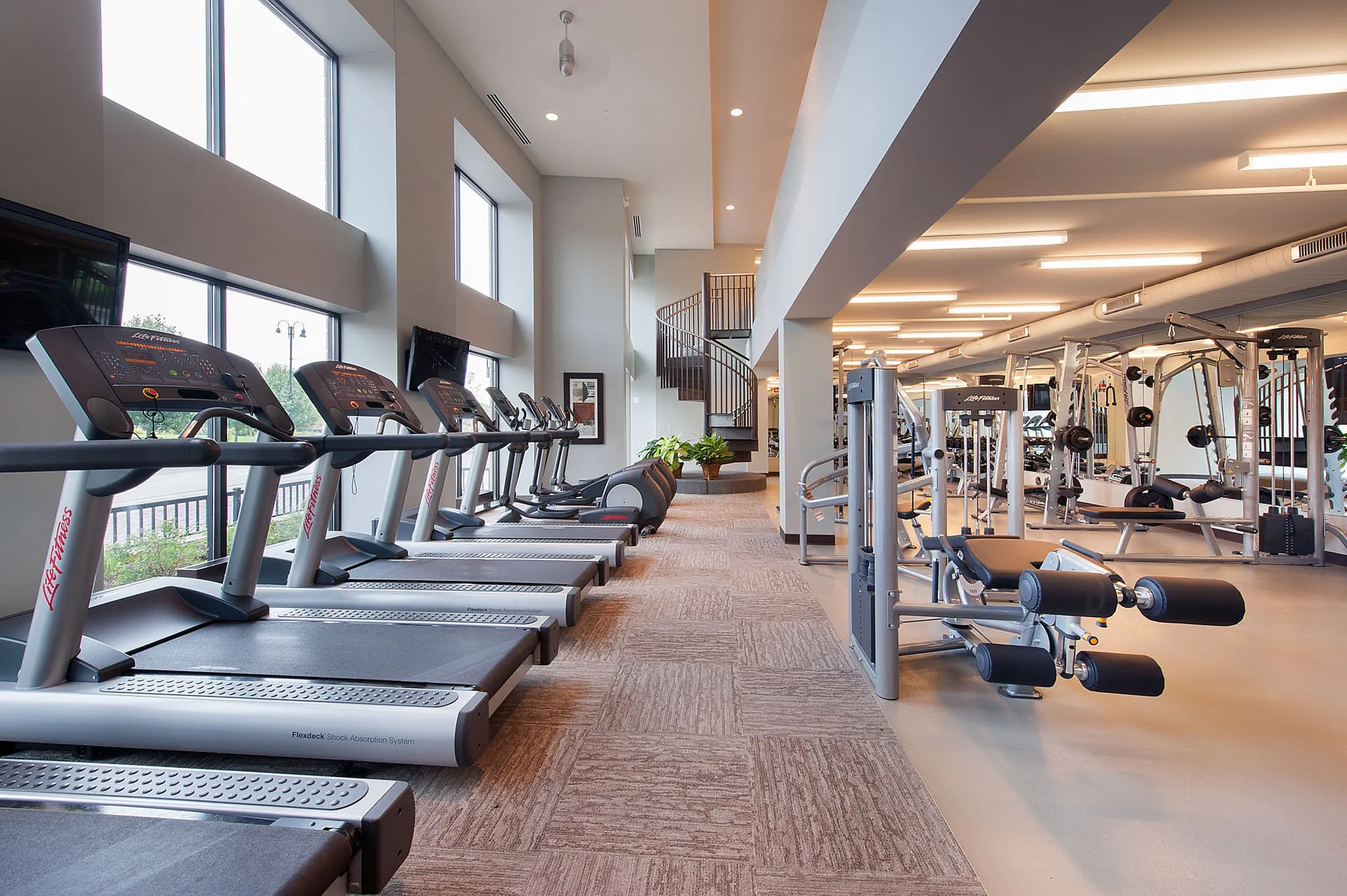 Bright and modern fitness center with state-of-the-art equipment, ample natural light, and a sleek spiral staircase.