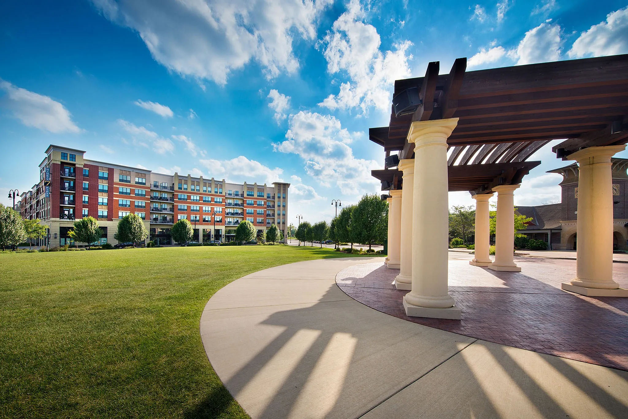 Elegant apartment complex with modern architecture, surrounded by lush green lawns and a stylish pergola under a bright blue sky.