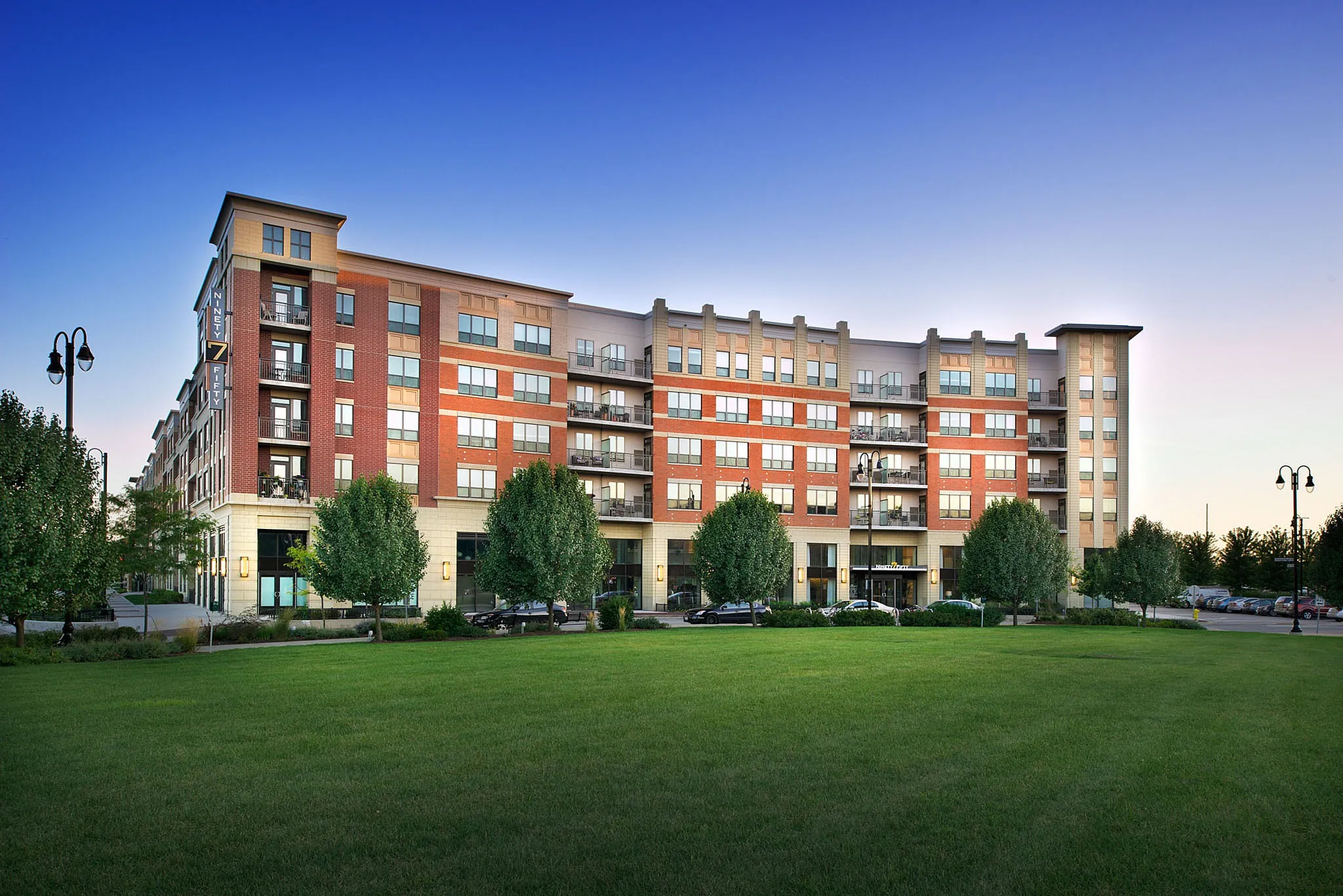 Contemporary apartment building with brick facade, expansive windows, and lush green landscaping under a clear blue sky.