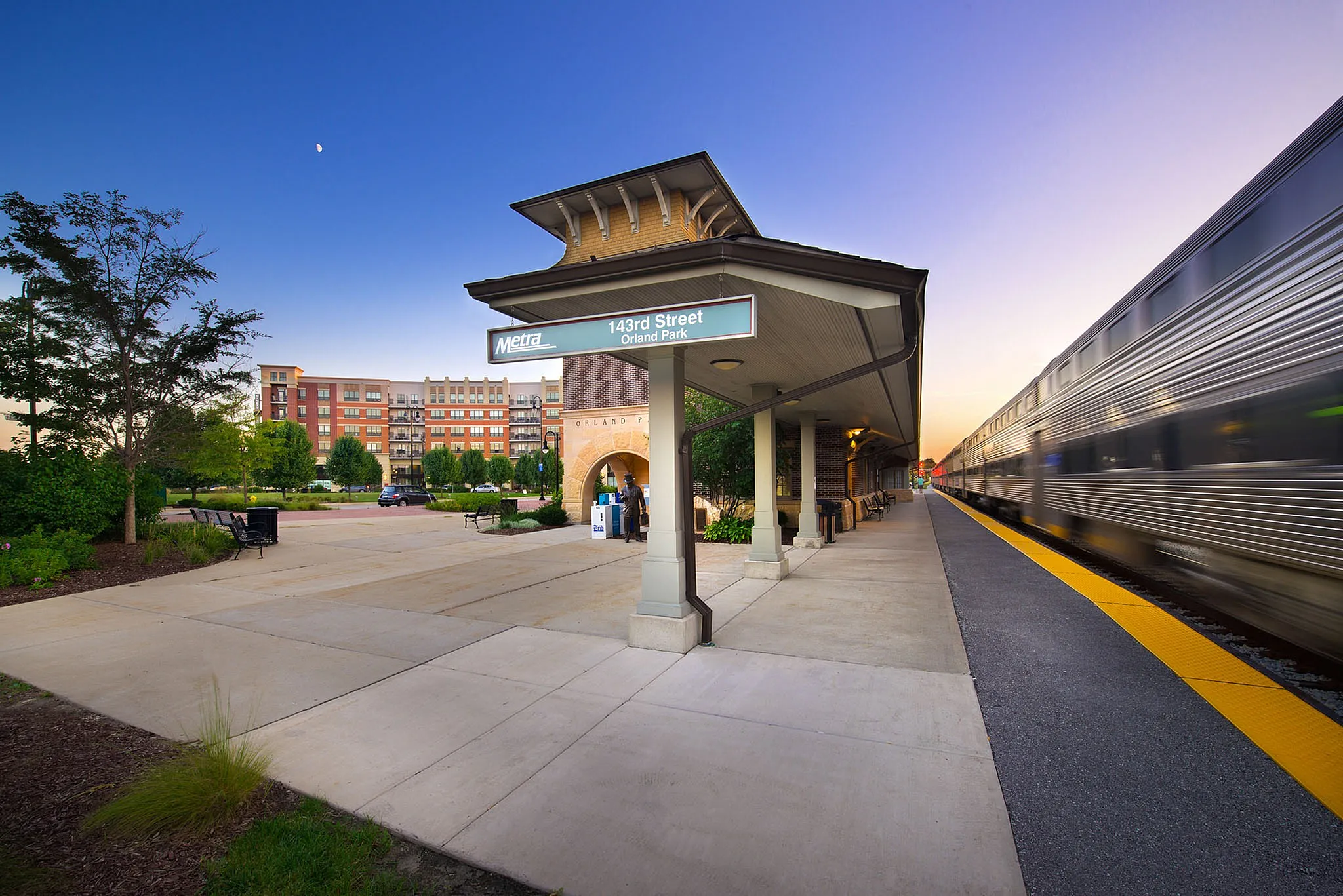 Charming 143rd Street Orland Park train station at dusk with sleek train passing, surrounded by lush greenery and modern buildings.