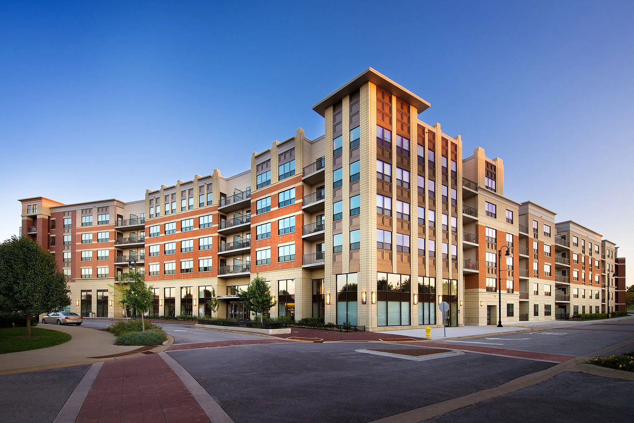 Modern apartment building with a brick facade, large windows, and private balconies, set in a beautifully landscaped urban area.