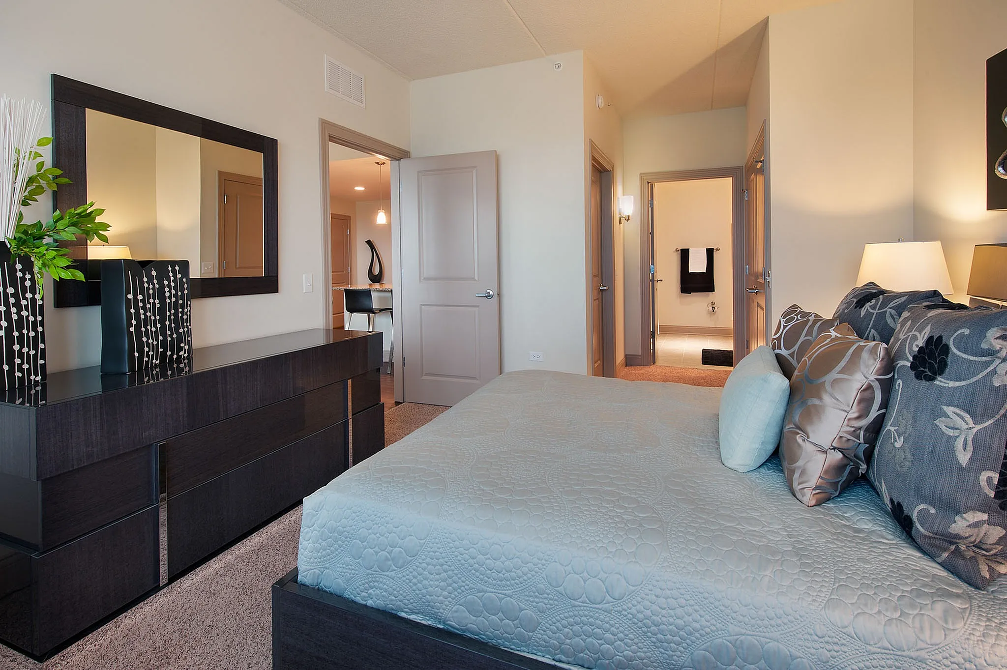 Elegant bedroom with plush bedding, modern dresser, and a glimpse into a stylish ensuite bathroom and cozy living area.