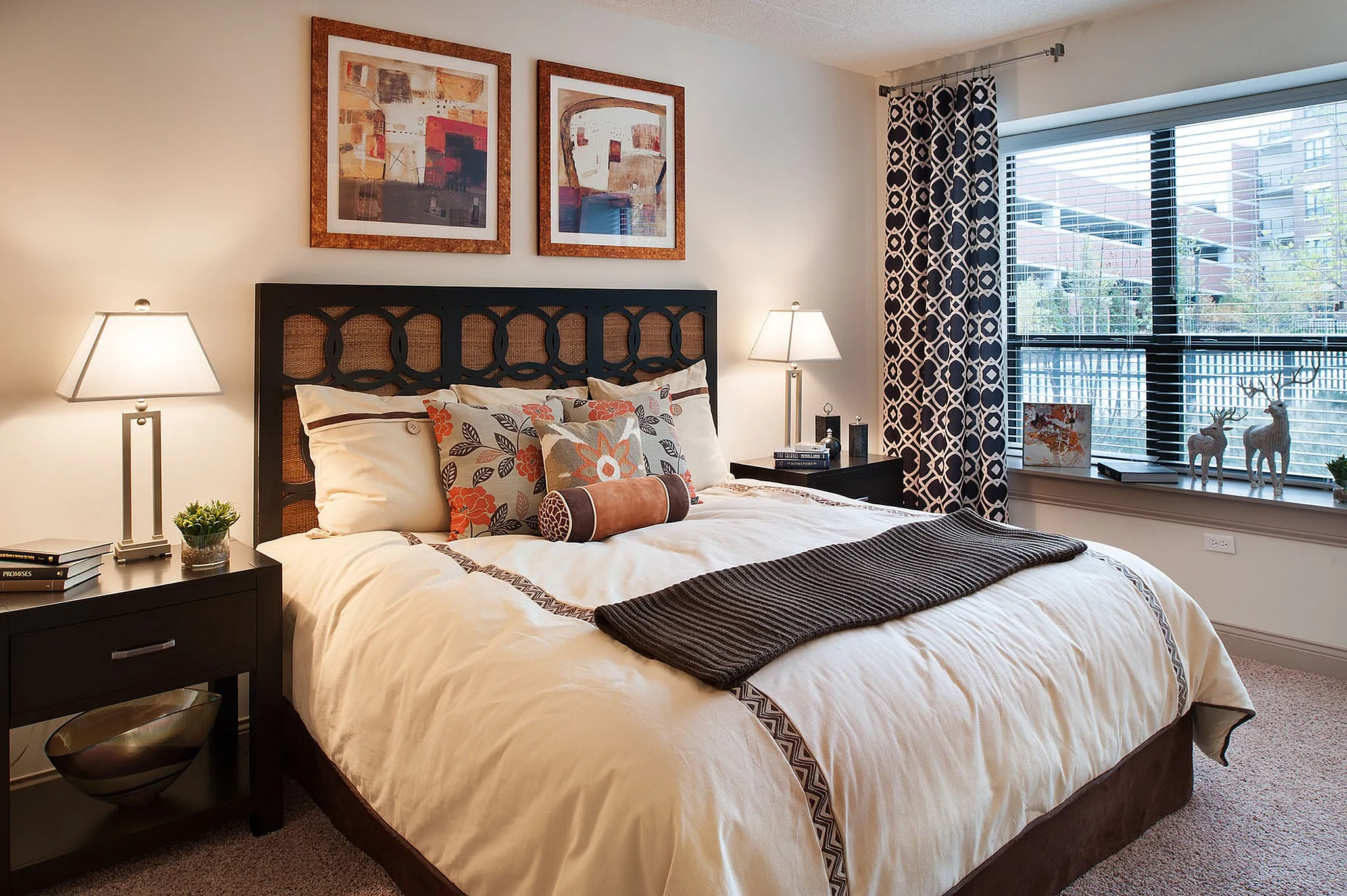 Inviting bedroom featuring a plush bed adorned with floral cushions, elegant bedside lamps, abstract wall art, and a large window with patterned curtains.