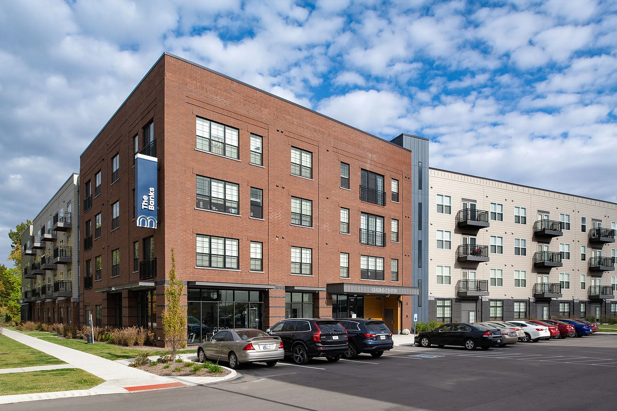 Modern apartment building with red brick facade, featuring large windows and private balconies, surrounded by lush greenery and ample parking space.