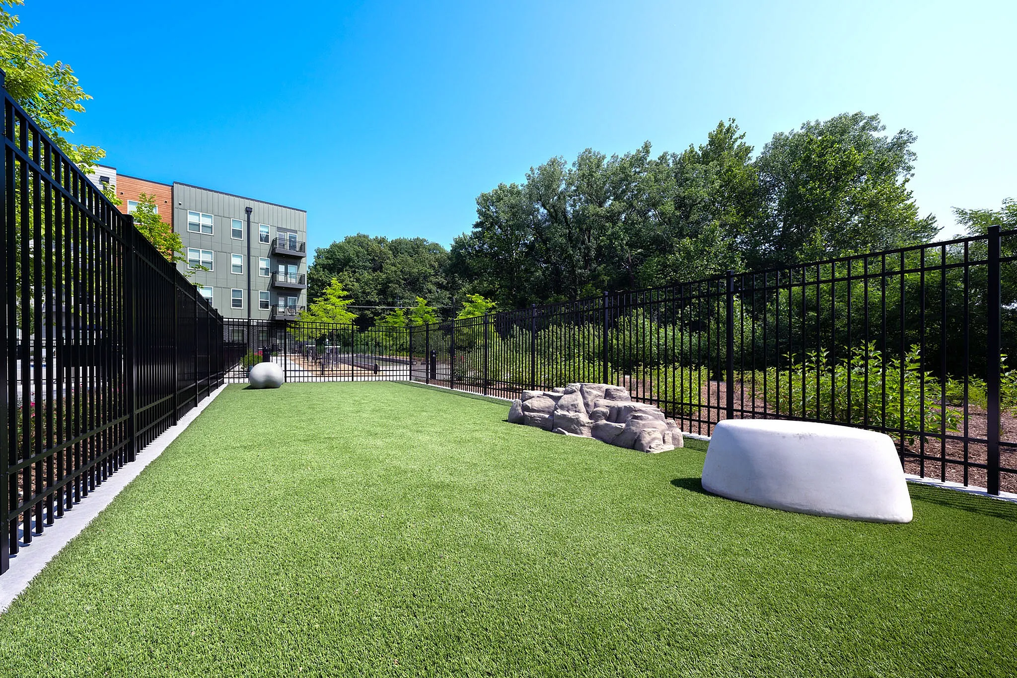 Modern apartment building with charming outdoor recreation area, lush greenery, and stylish fencing under a clear blue sky.