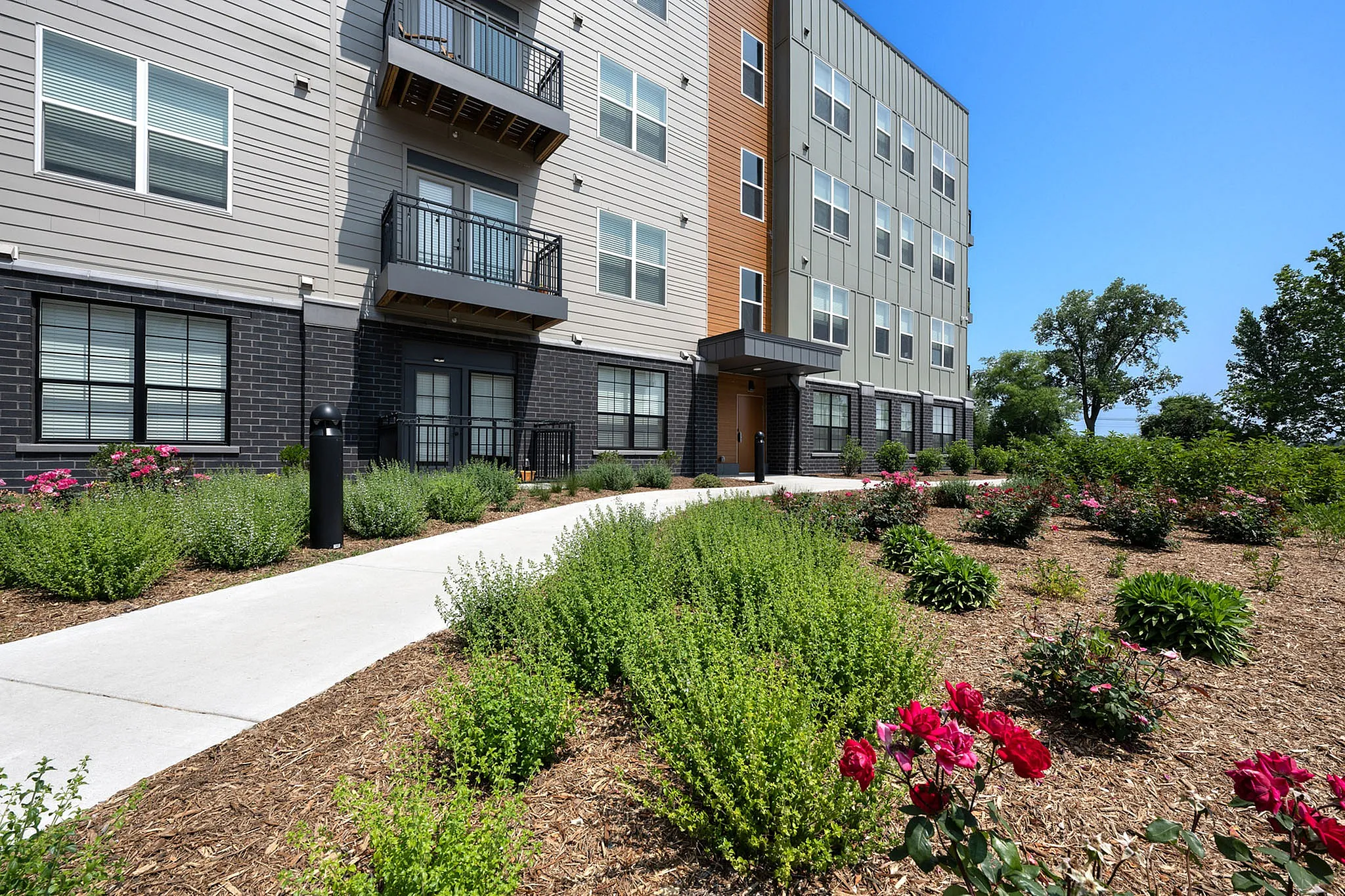 Modern apartment complex with vibrant landscaping, featuring lush greenery and colorful flowers, set against a clear blue sky.