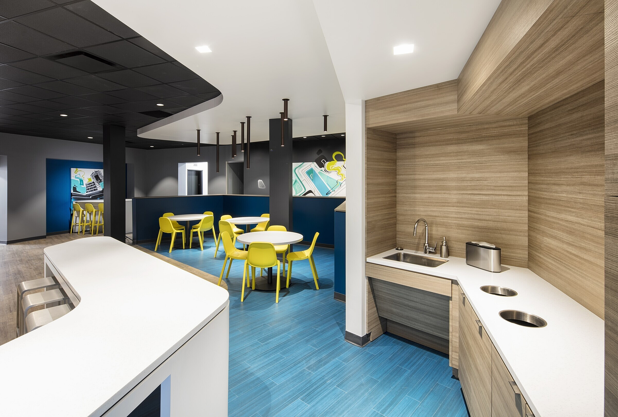 Modern common area with vibrant yellow chairs, stylish blue flooring, and a compact wood-paneled kitchenette.