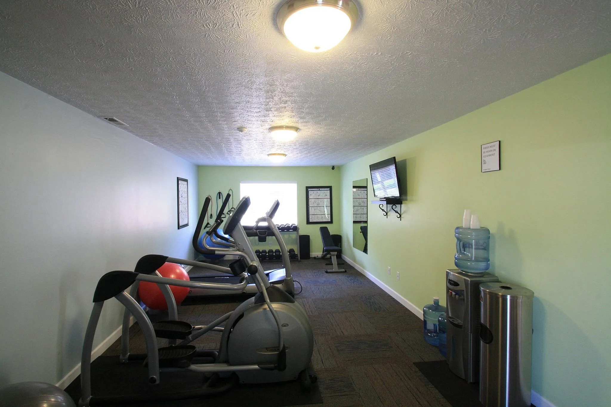 Inviting fitness center with modern exercise equipment, bright green walls, and cozy atmosphere, complete with a water cooler and entertainment screen.