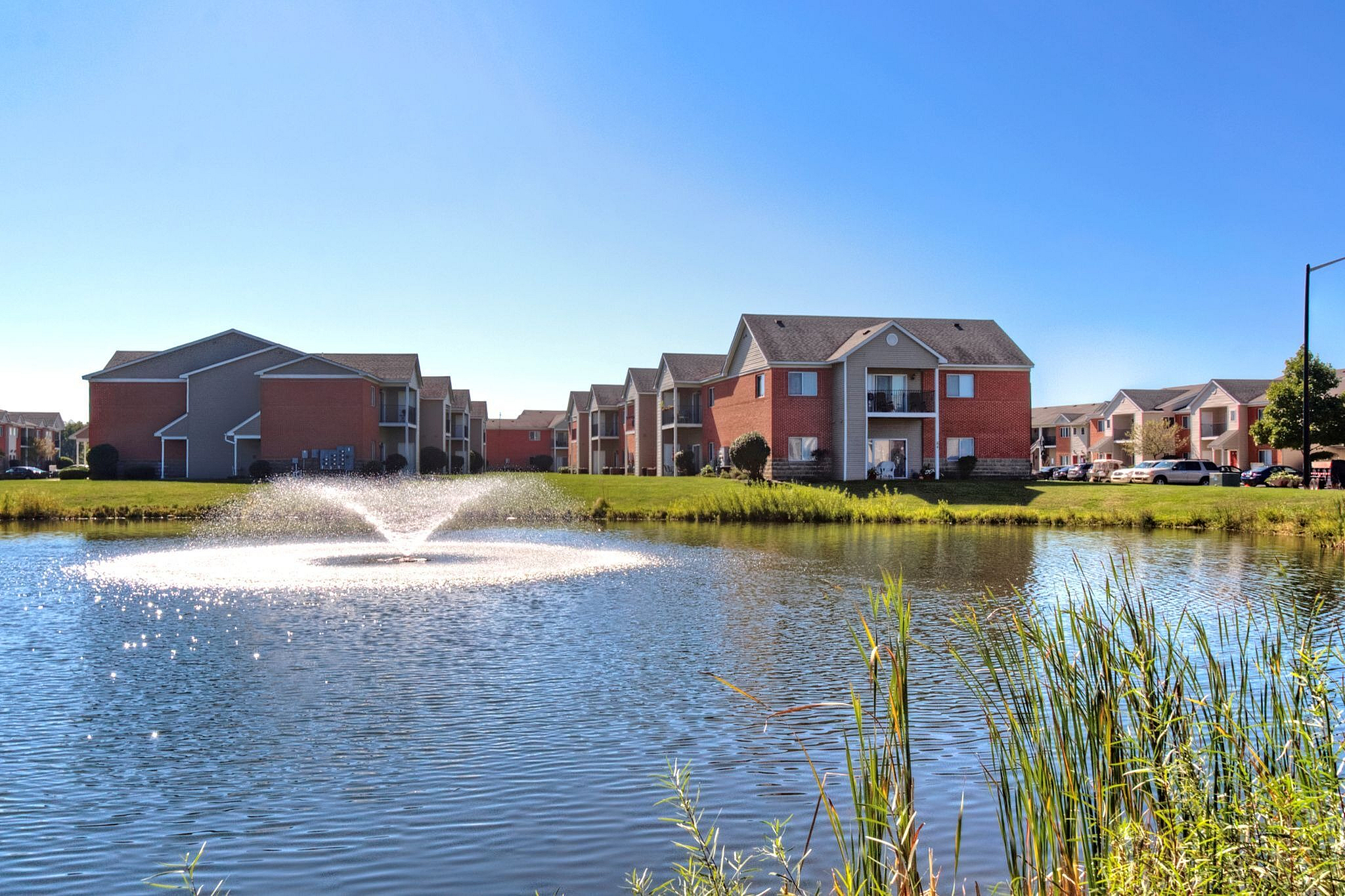 Charming apartment complex with picturesque pond and fountain views, surrounded by lush greenery under a clear blue sky.