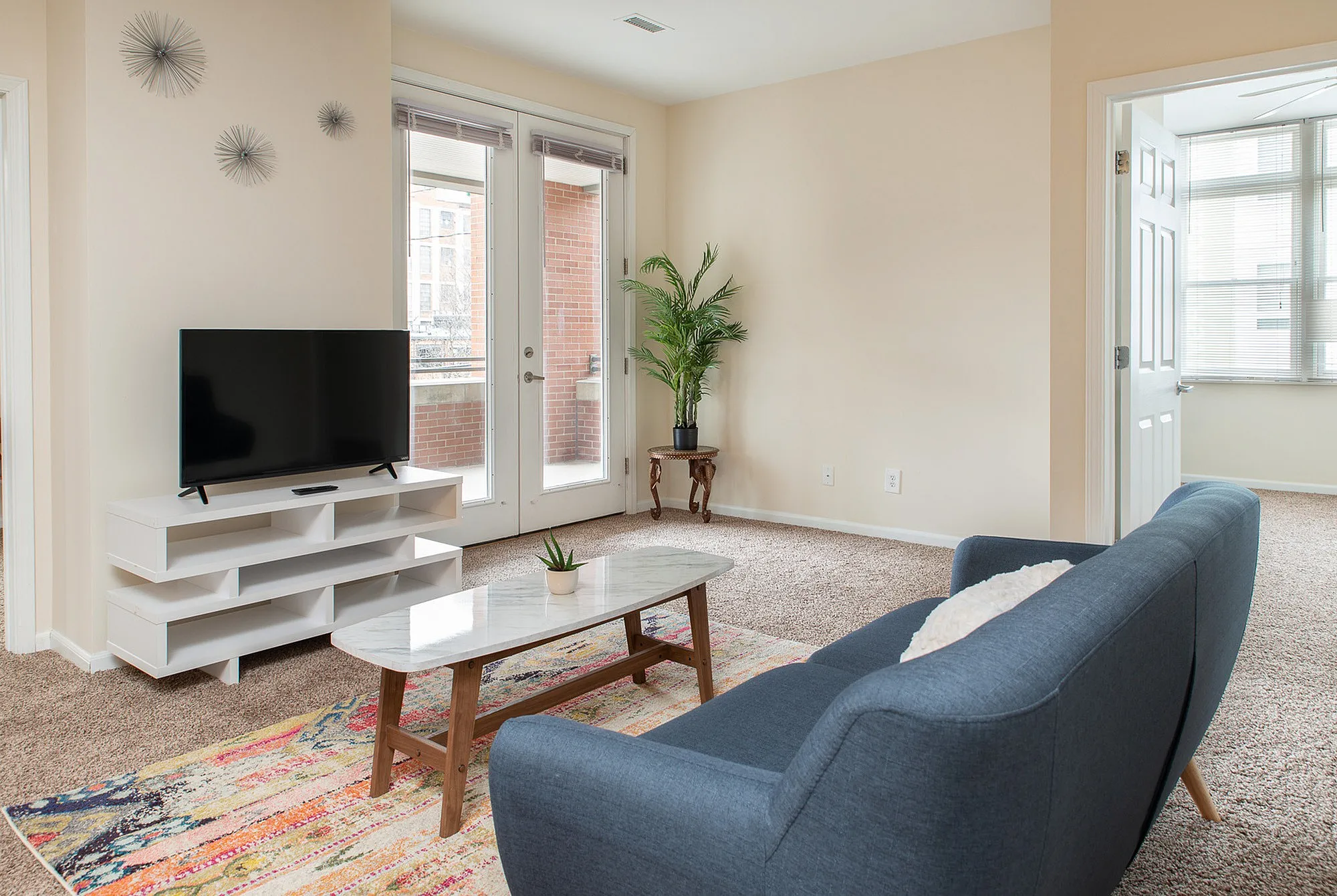 Bright and inviting living room with modern decor, featuring a sleek TV stand, stylish blue sofa, and a lush potted plant, illuminated by natural light from glass doors leading to a balcony.